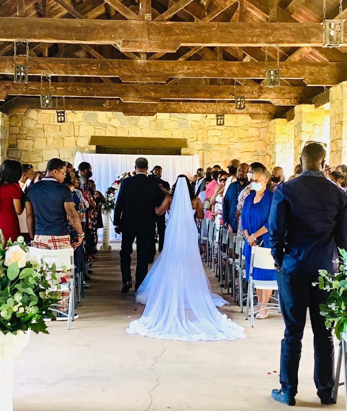 Bride walking down the aisle under a rustic wooden pavilion with stone walls.