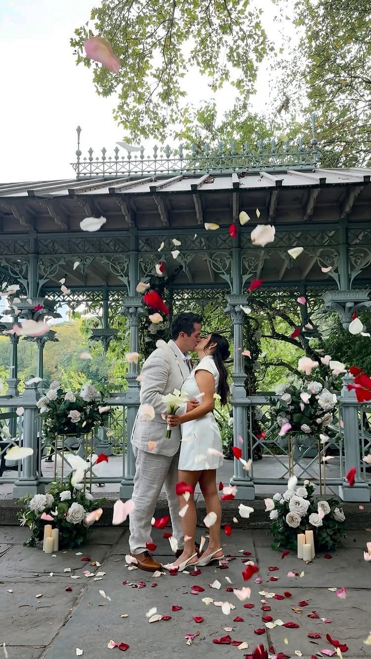 Couple kissing under an ornate metal gazebo with falling flower petals in a park