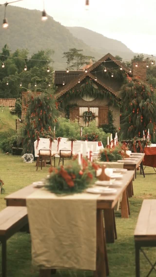 Wooden wedding tables with greenery centerpieces on a lawn before a rustic cottage.