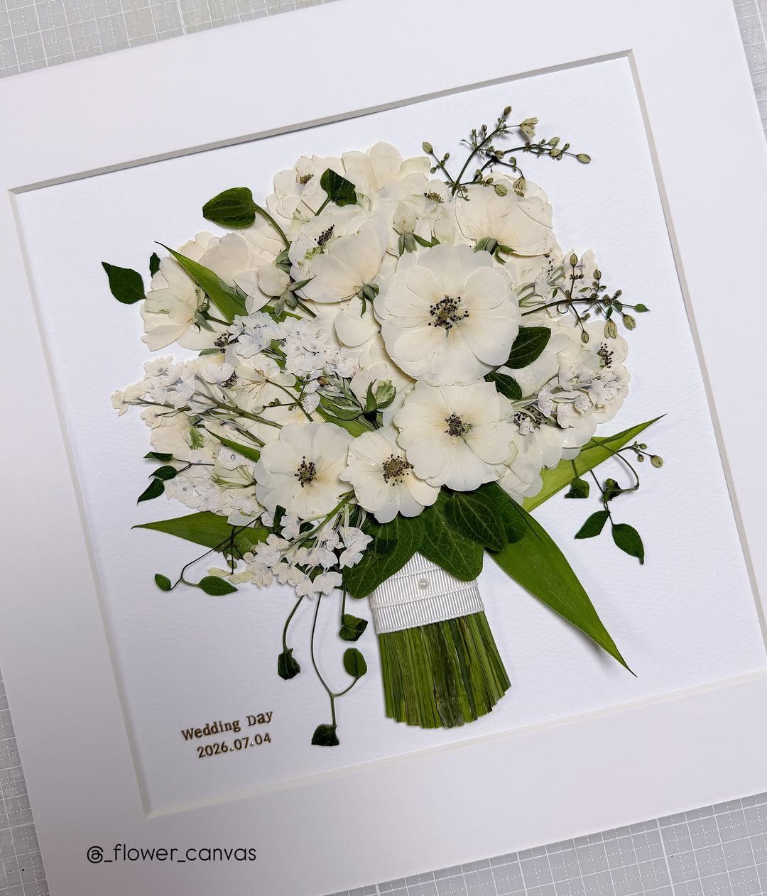Pressed white wedding bouquet with green leaves framed in a white mat.