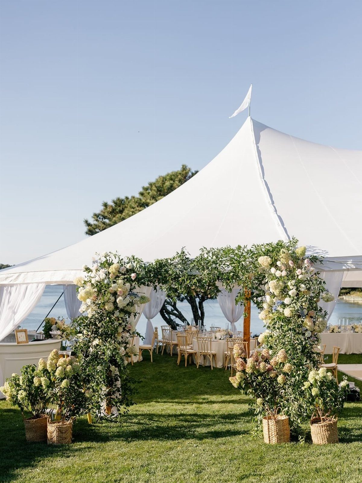 Floral archway leading into a white wedding tent on a waterfront lawn.