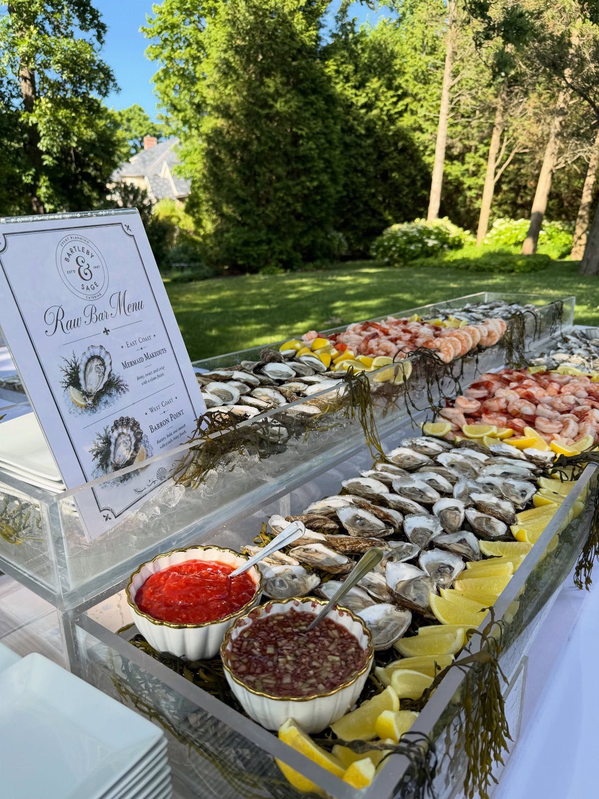 Seafood raw bar display with oysters and shrimp on clear acrylic tiers outdoors.