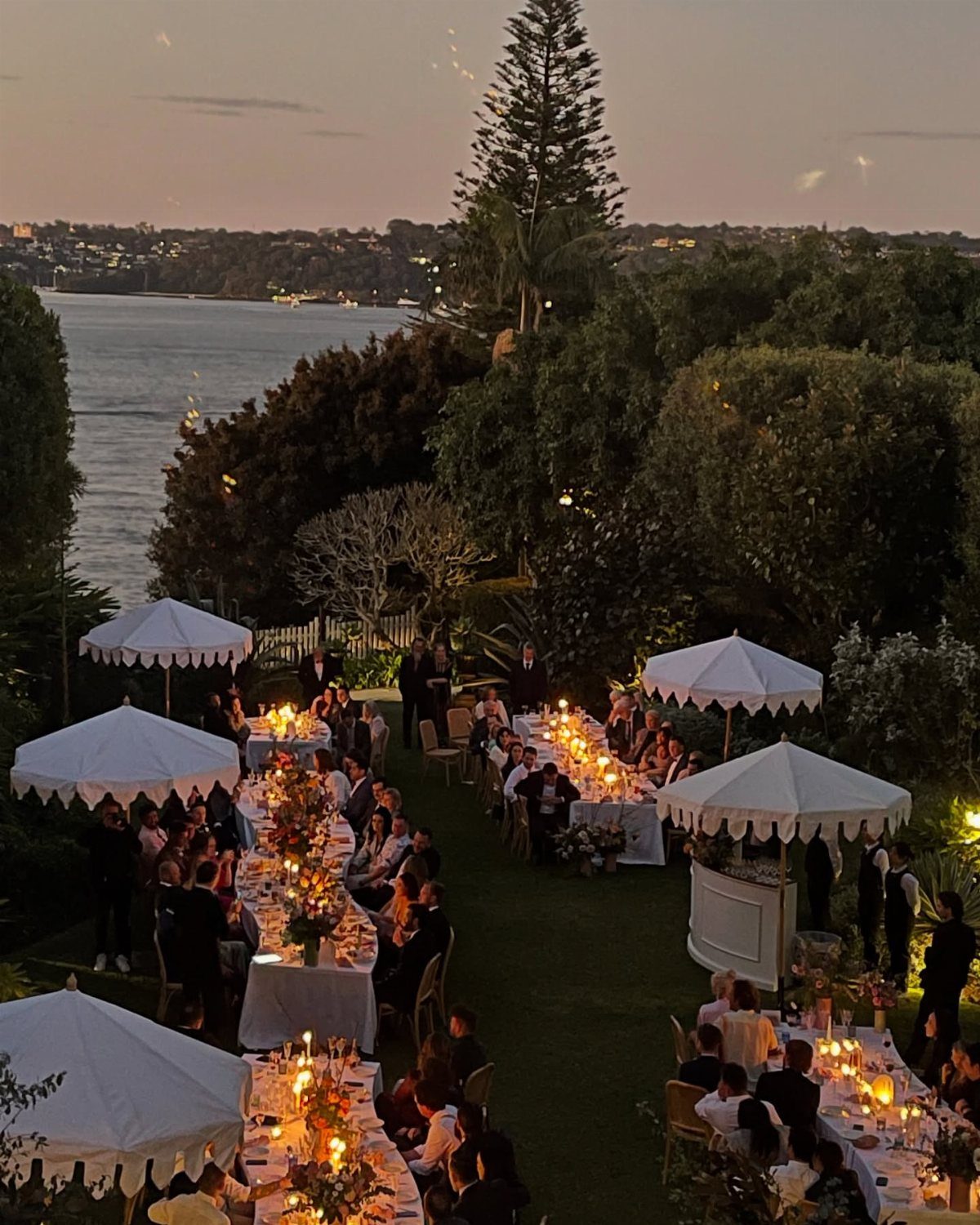 Outdoor wedding reception with long candlelit tables on a lawn overlooking a bay.