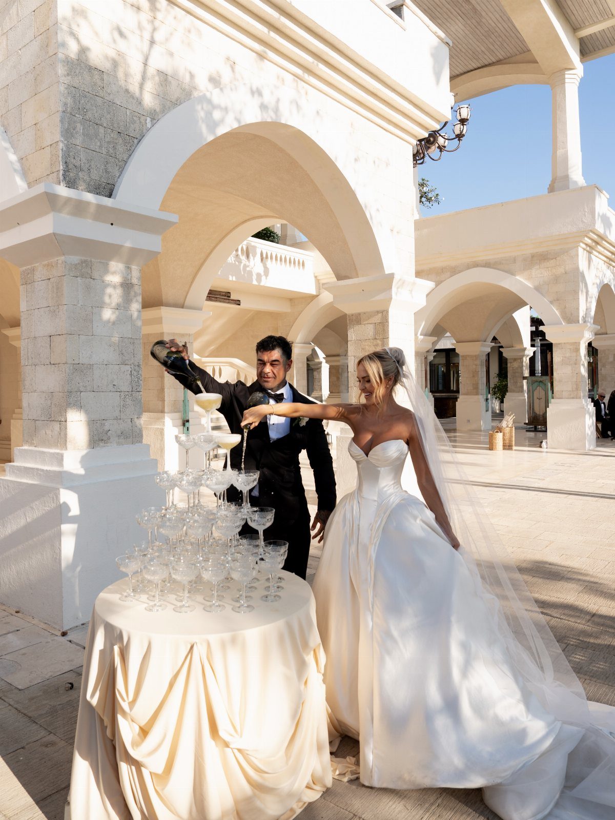 Bride and groom pouring champagne into a glass tower at a wedding celebration.