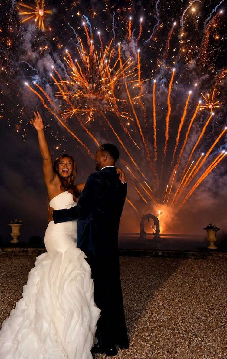 Bride and groom embracing under a sky filled with exploding orange fireworks.