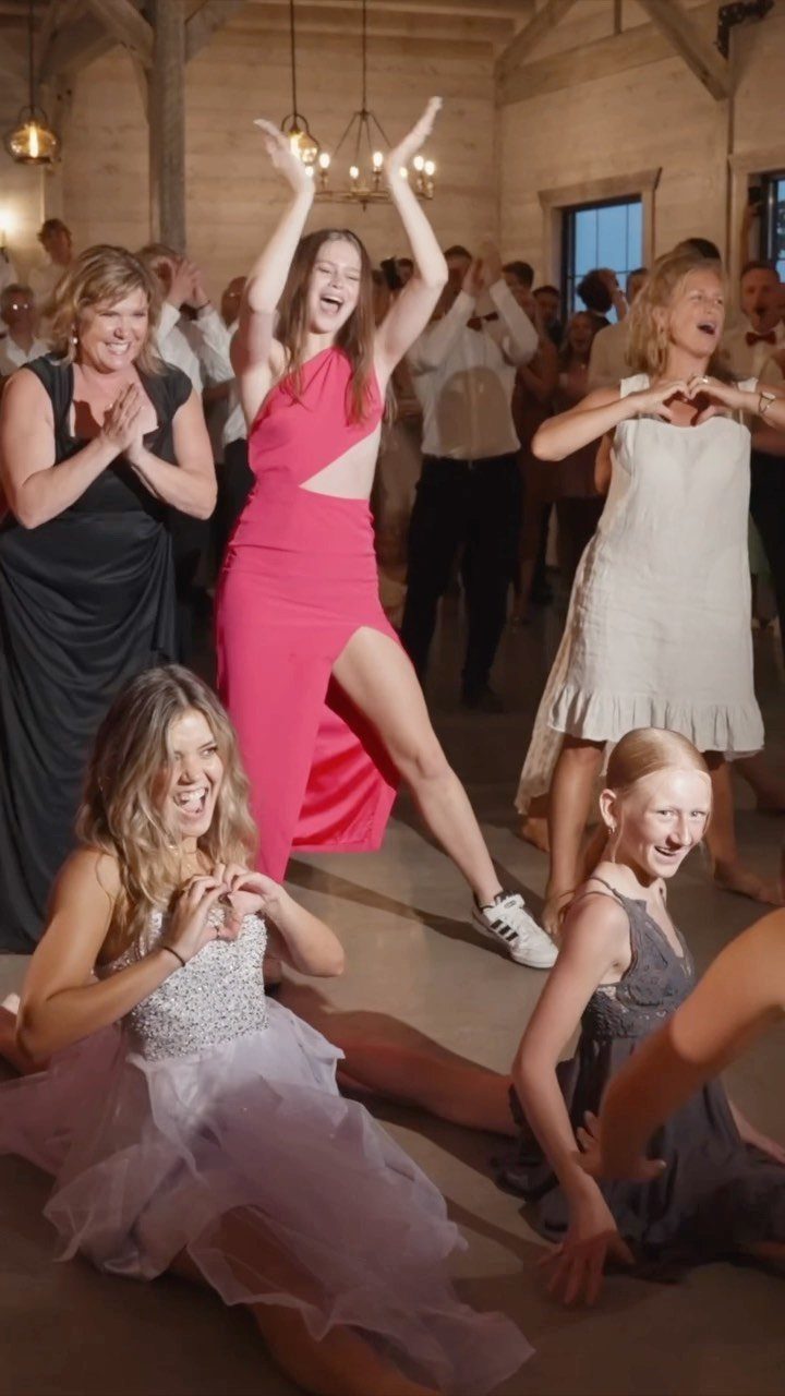 Women in formal dresses dancing and laughing together in a rustic wedding venue.