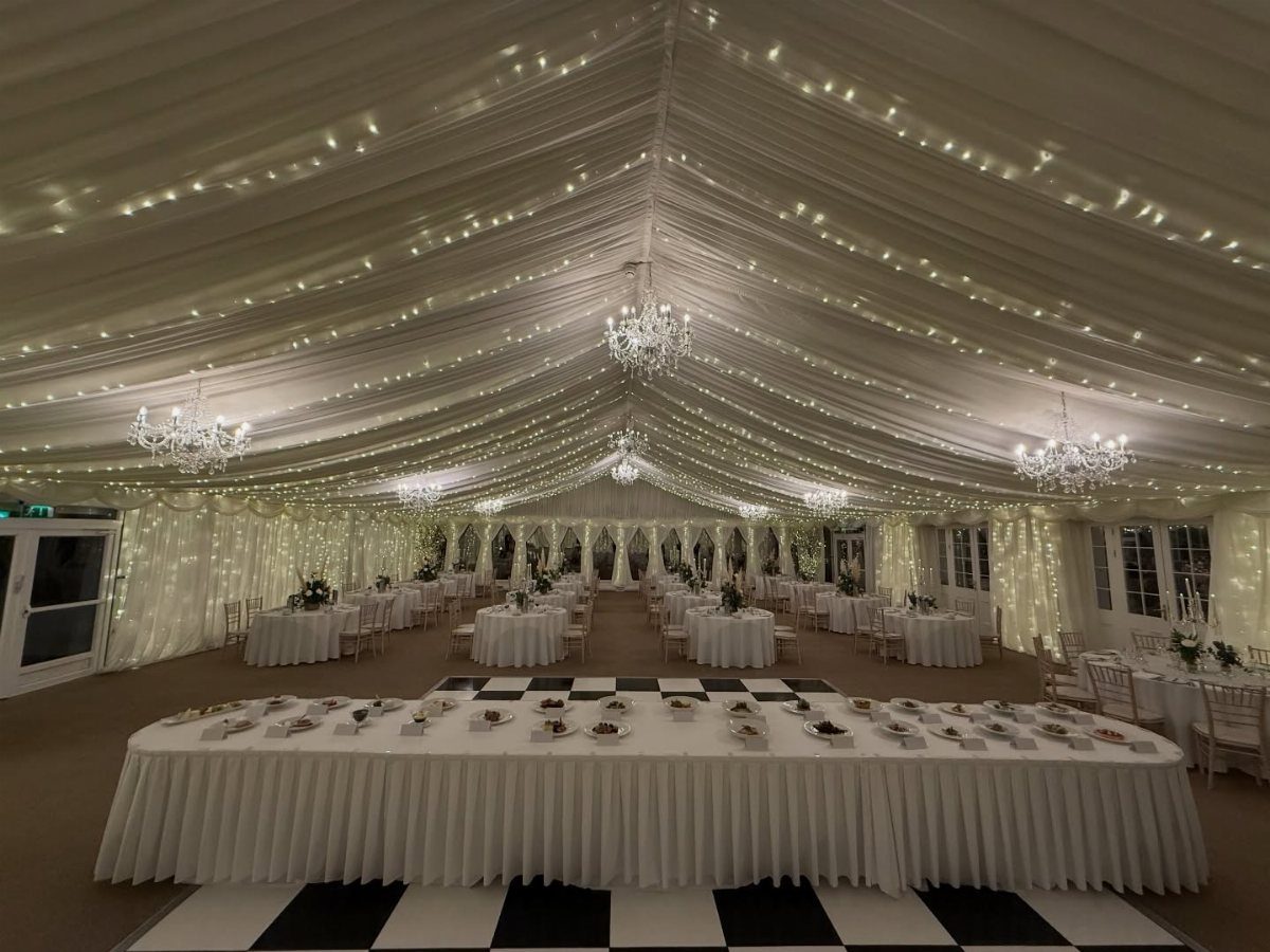 White event tent with draped ceiling, fairy lights, chandeliers, and set dining tables.