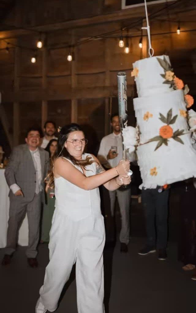 Woman in white jumpsuit hitting a cake-shaped piñata at a wedding celebration.