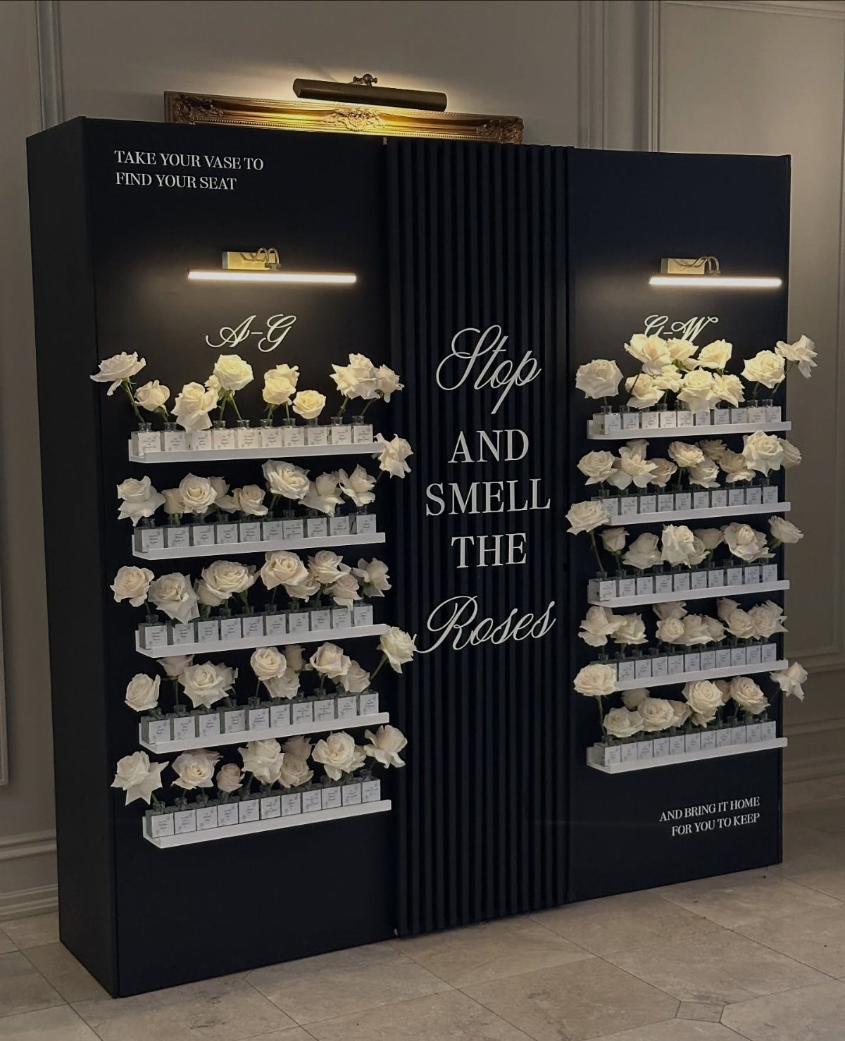 Black seating chart wall with white roses in vases and gold lighting.