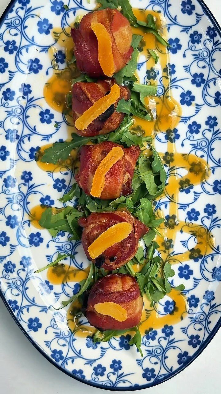 Bacon-wrapped scallops with orange slices on arugula over a blue floral platter.