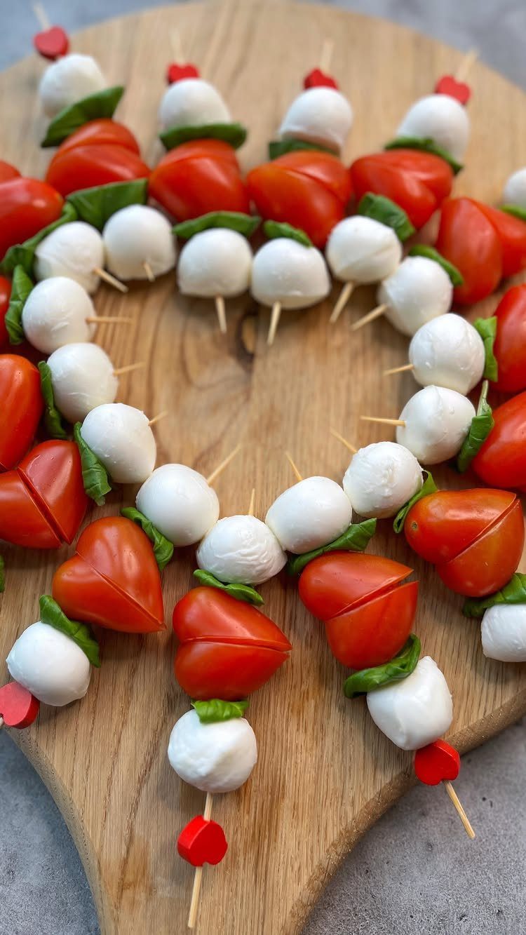 Heart-shaped Caprese salad skewers with tomatoes, mozzarella, and basil on a wooden board.