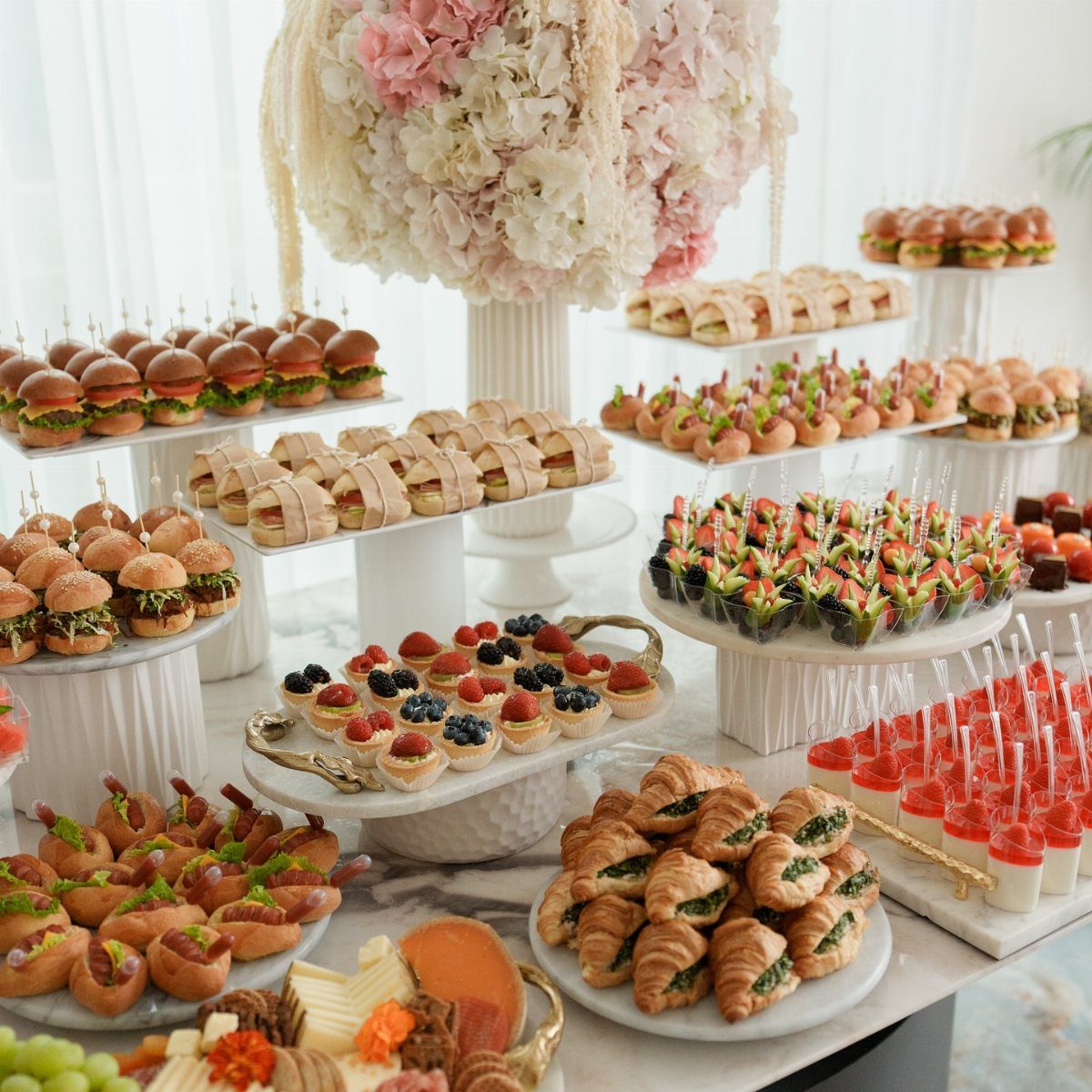 Wedding appetizer spread with mini sliders, sandwiches, and fruit tarts on marble tables.