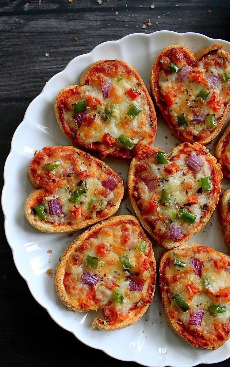 Heart-shaped mini pizzas with melted cheese and vegetables on a white scalloped plate.