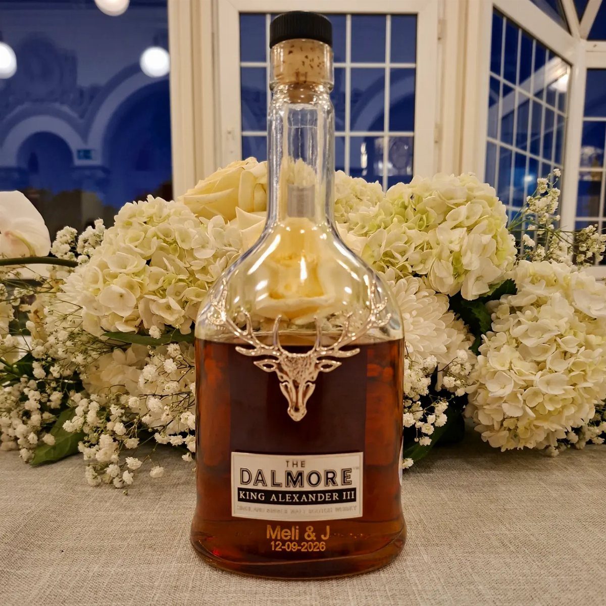 Whiskey bottle centerpiece with personalized label surrounded by white hydrangea floral arrangements.