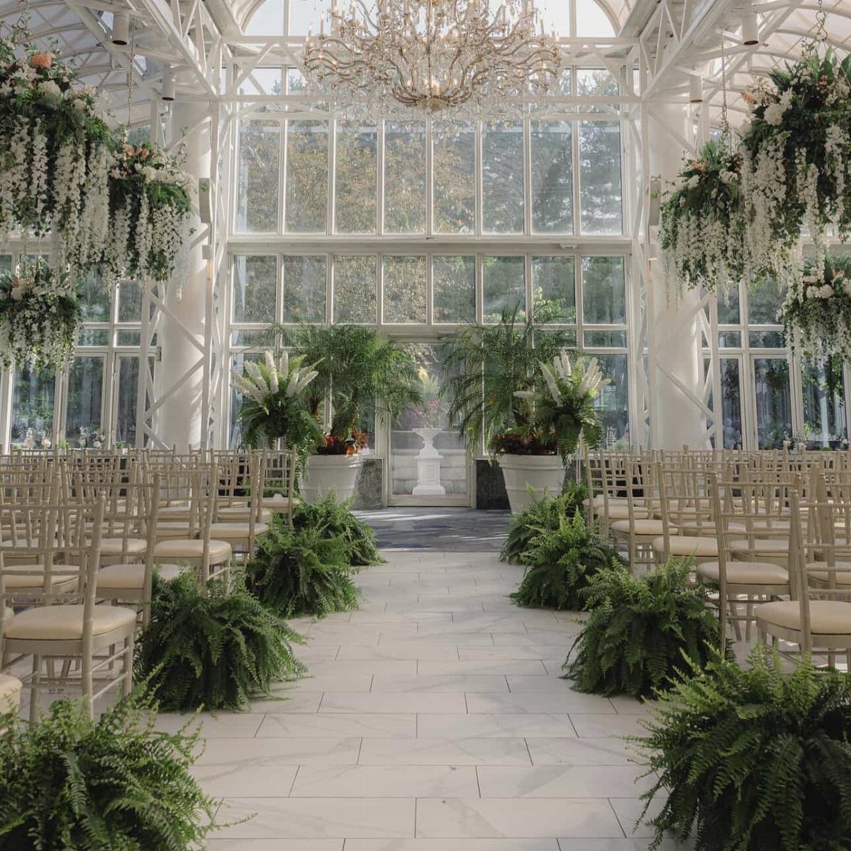 White glass conservatory wedding venue with marble floors, beige chairs, and lush greenery.