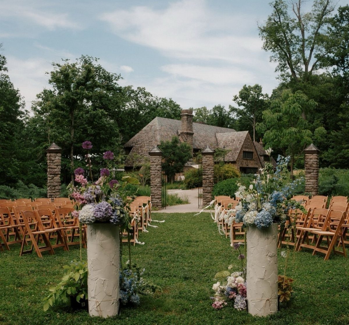 Outdoor wedding ceremony setup with wooden chairs and floral pedestals before a rustic cottage.