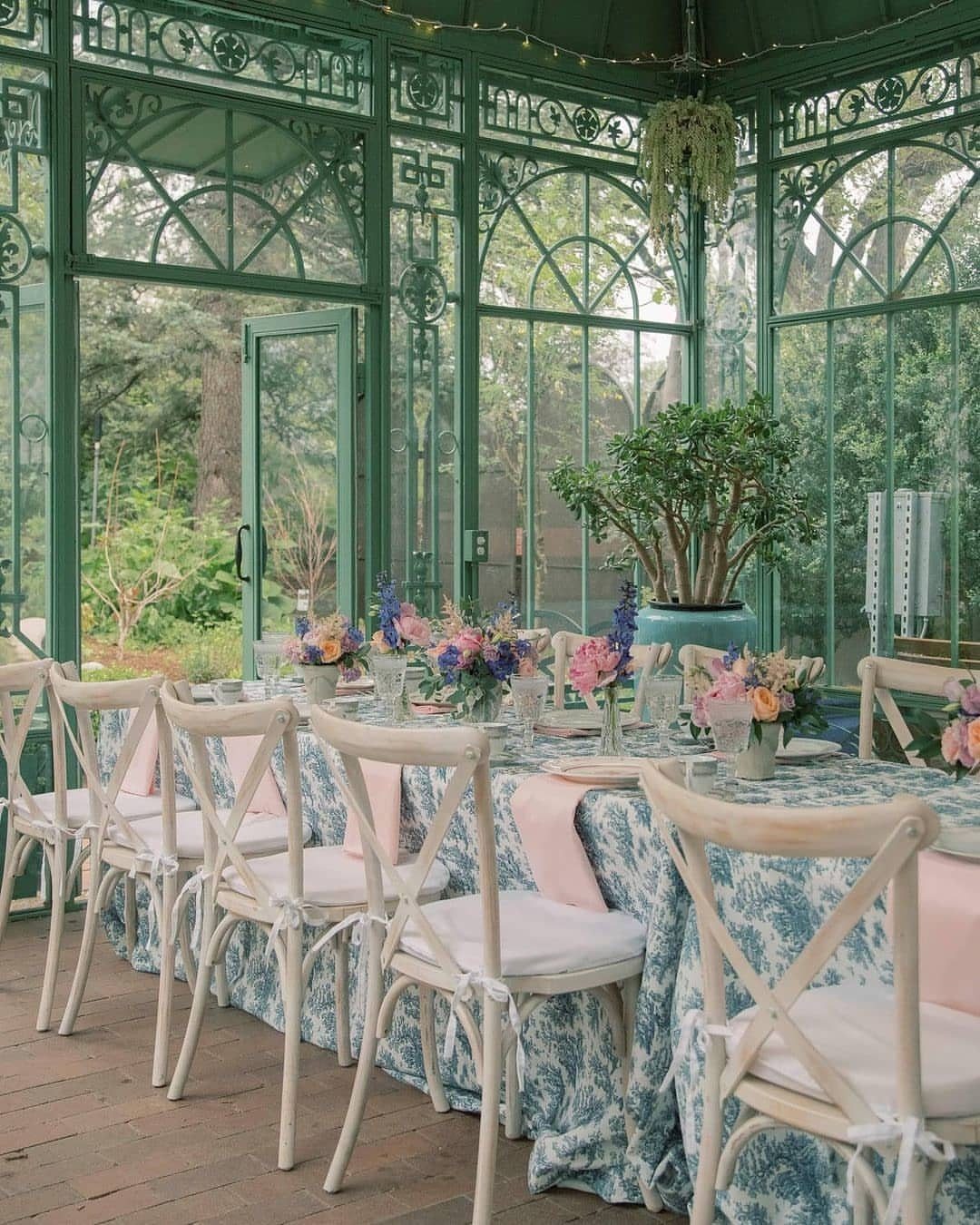 Long banquet table with floral centerpieces inside a green glass garden conservatory.