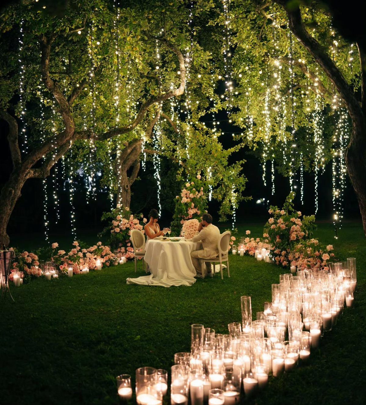 Couple dining under a tree draped in hanging string lights and candlelit paths.