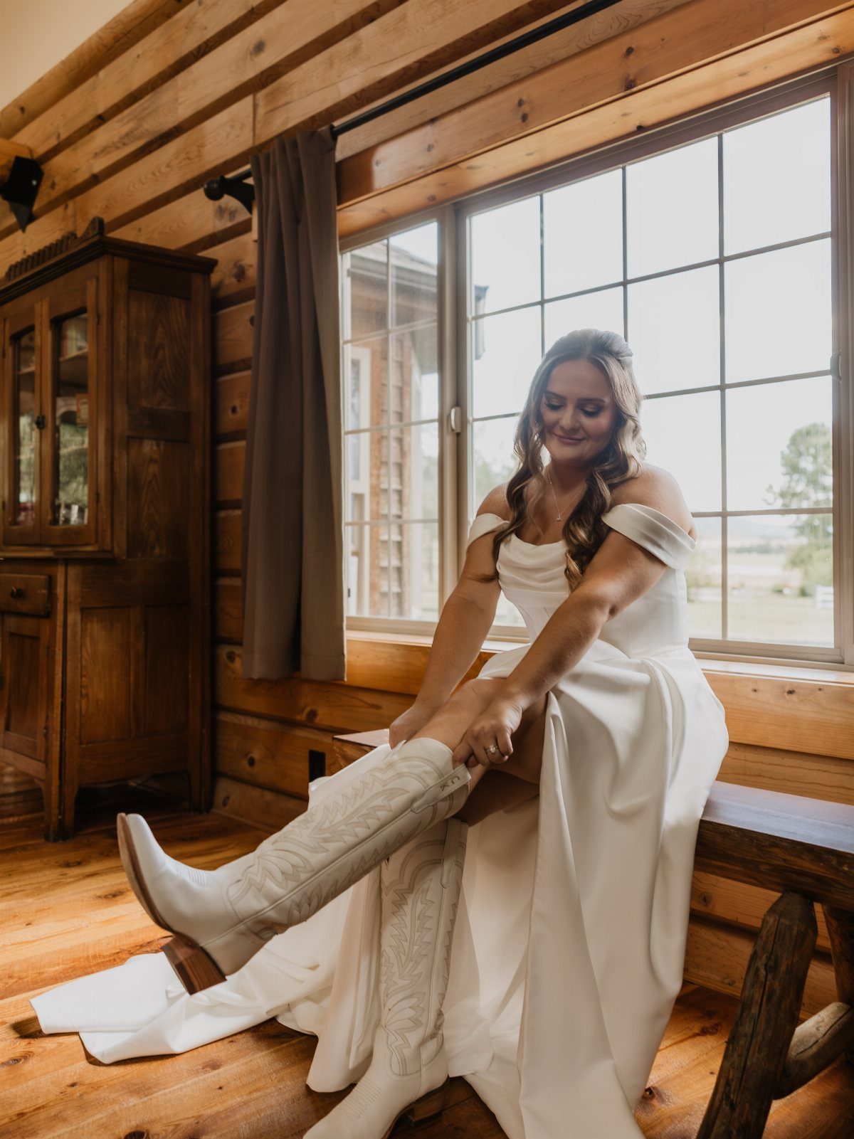 Bride in white gown putting on white cowboy boots inside a log cabin.