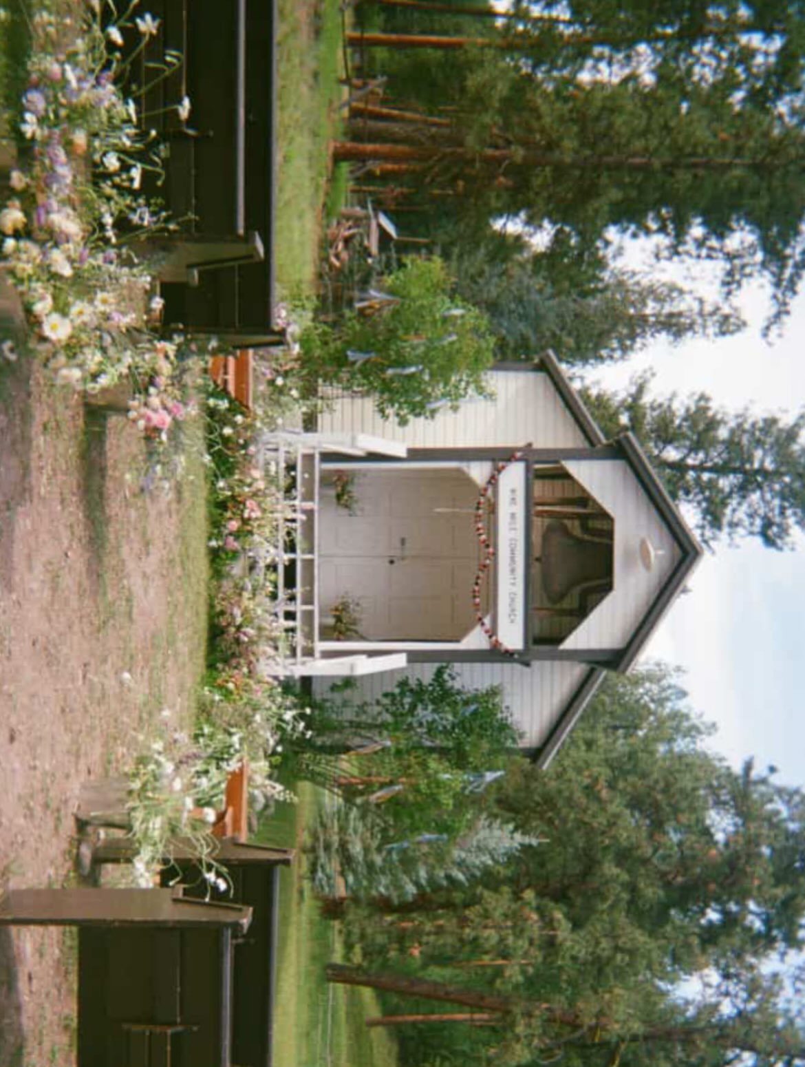White wooden chapel decorated with flowers in a pine forest with wooden benches.