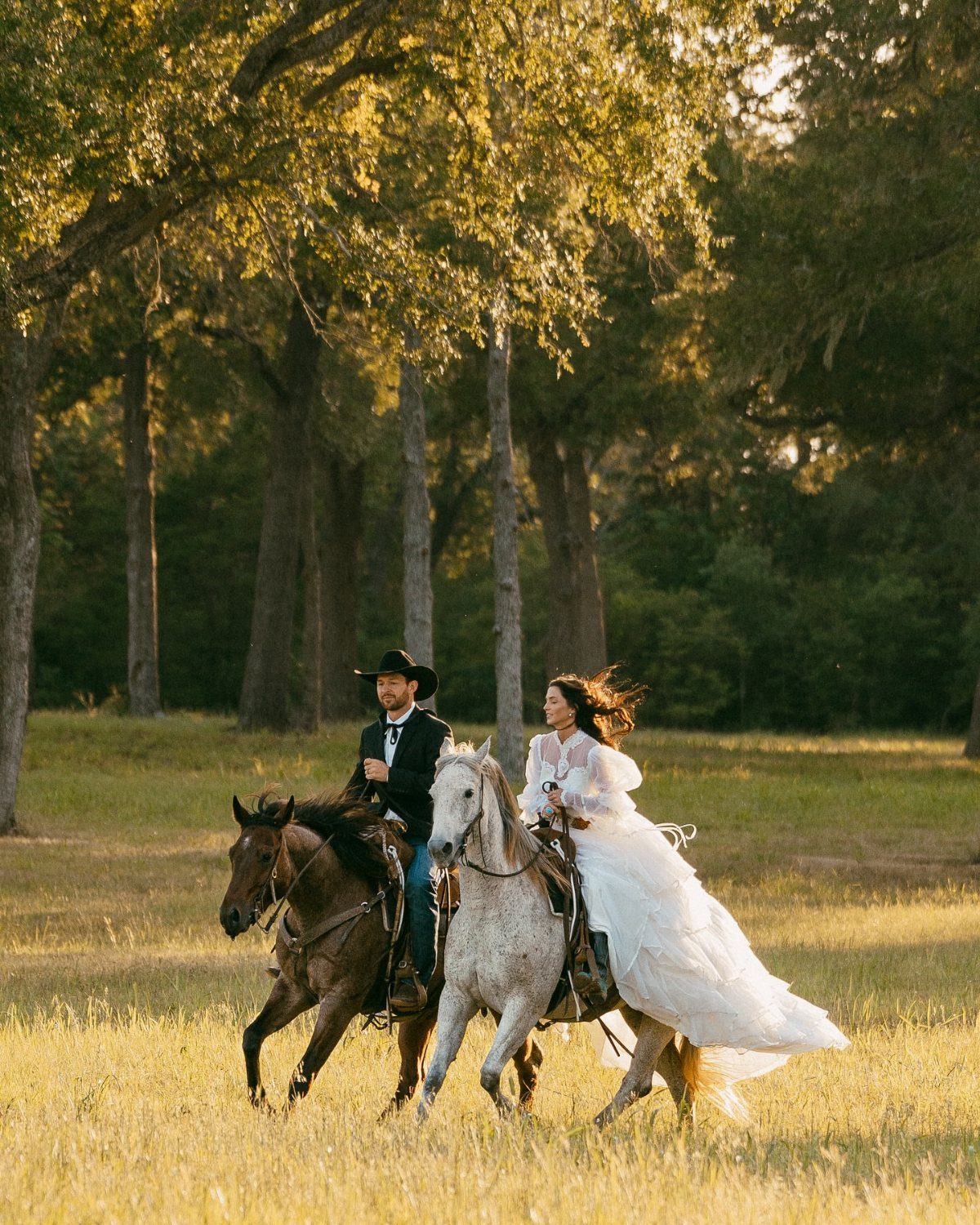 Bride and groom riding horses through a sunlit field with tall trees.