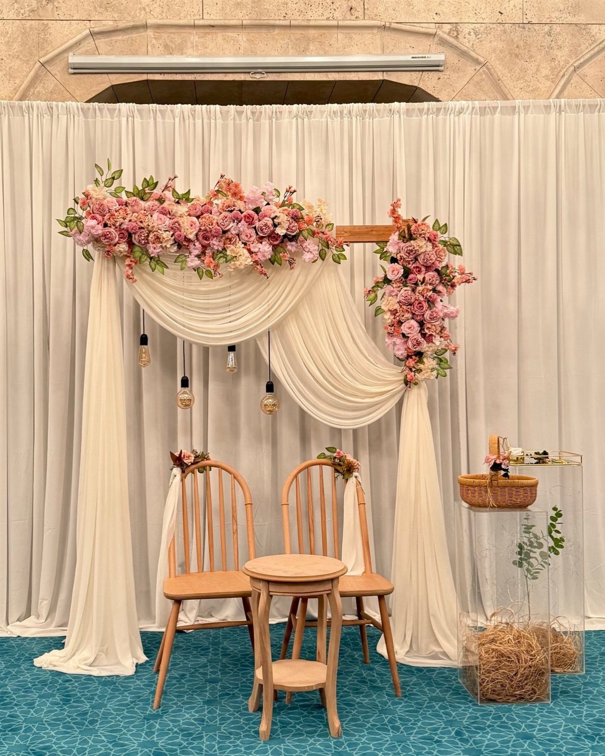 Floral wedding arch with pink roses and white draping behind two wooden chairs.