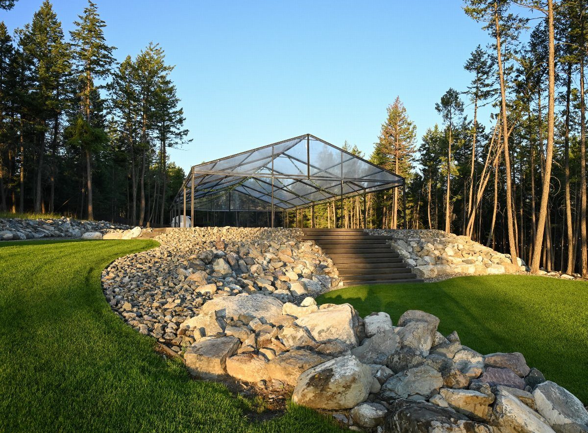 Modern glass pavilion on a rocky hill surrounded by tall pine trees and grass.