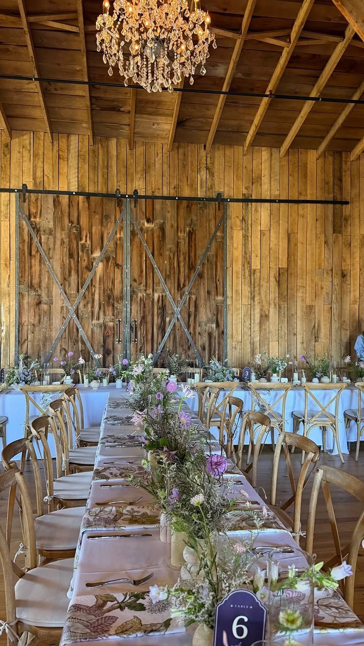 Long dining table with floral arrangements and crystal chandelier in a rustic barn.