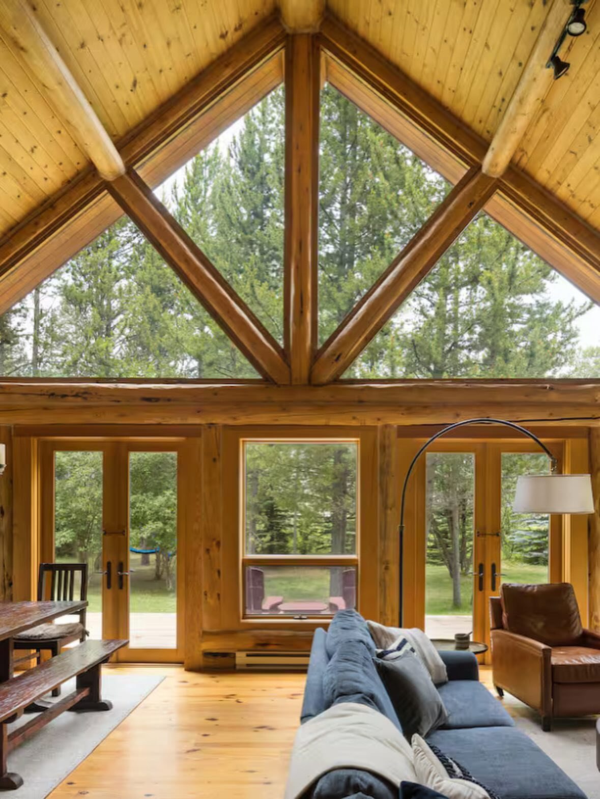Rustic log cabin interior with large windows overlooking a dense pine forest.