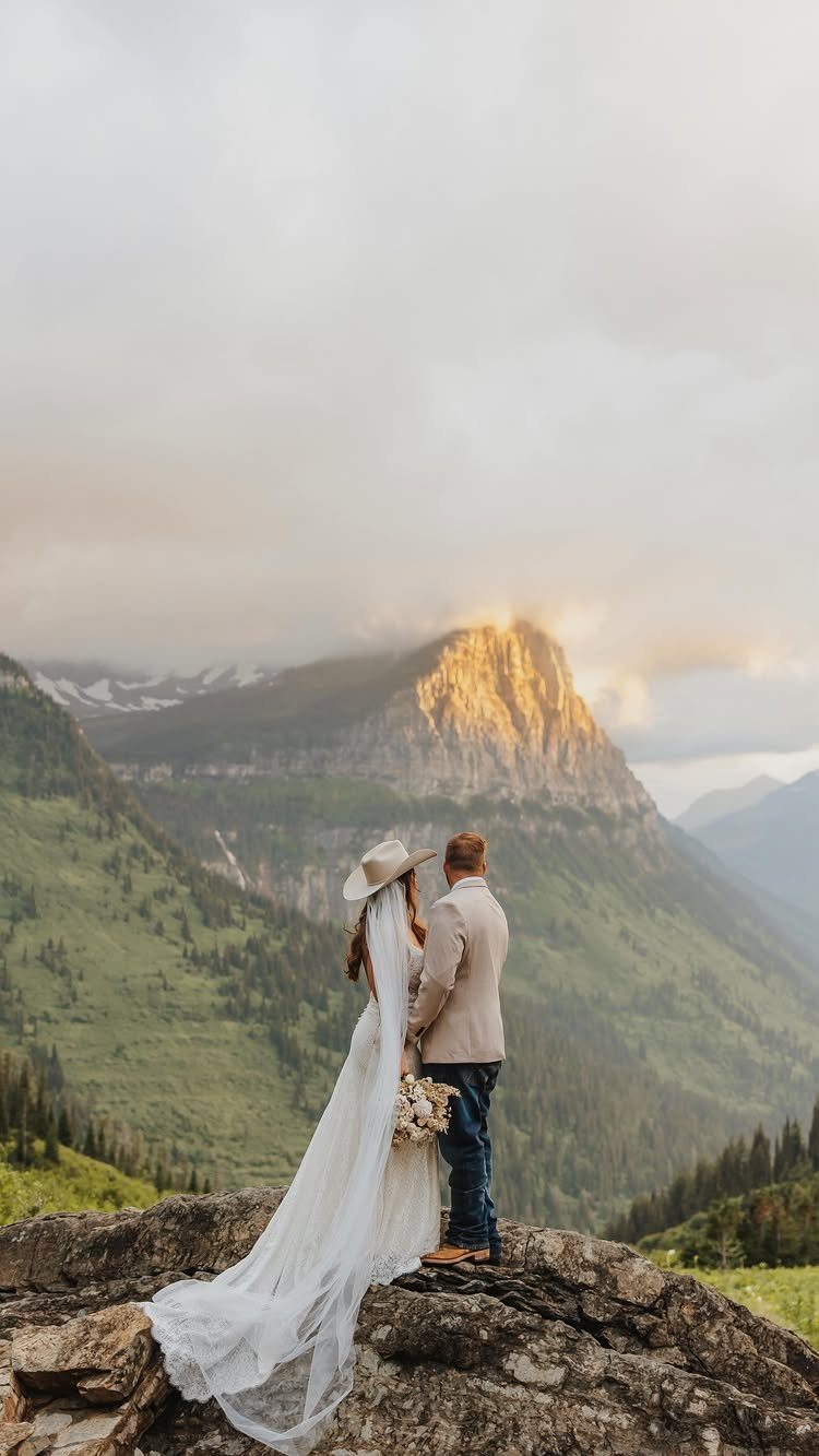 Bride and groom in western attire standing on a cliff overlooking Montana mountains.