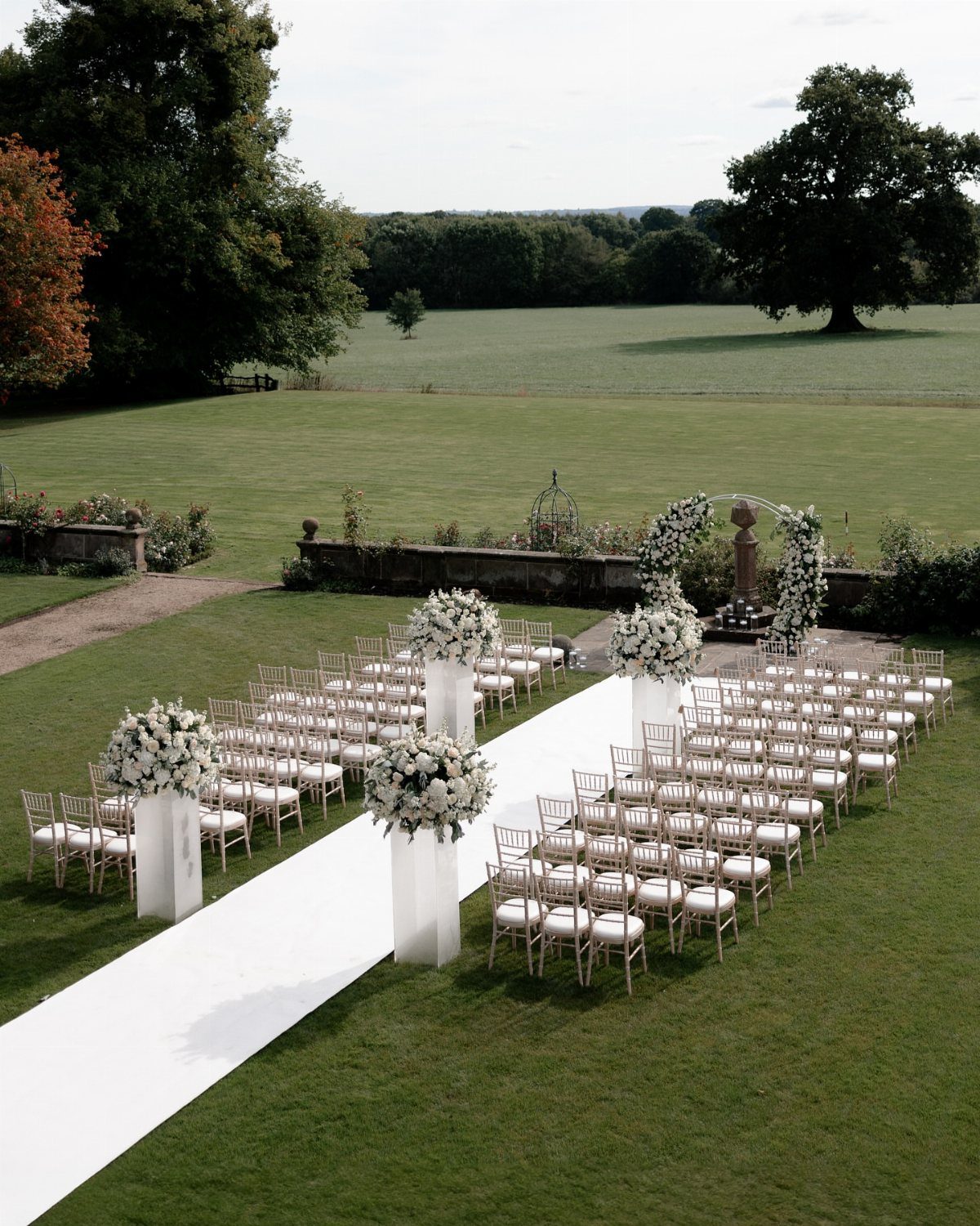 Outdoor wedding ceremony setup with white aisle, gold chairs, and floral arrangements.