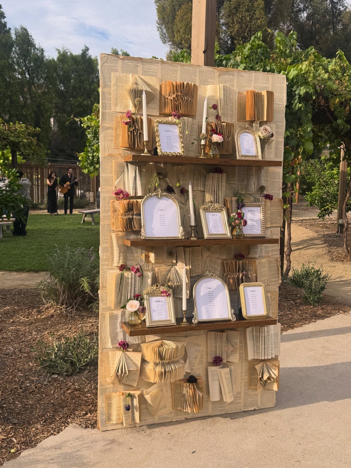 Rustic wooden seating plan board with gold frames and paper bundles outdoors.