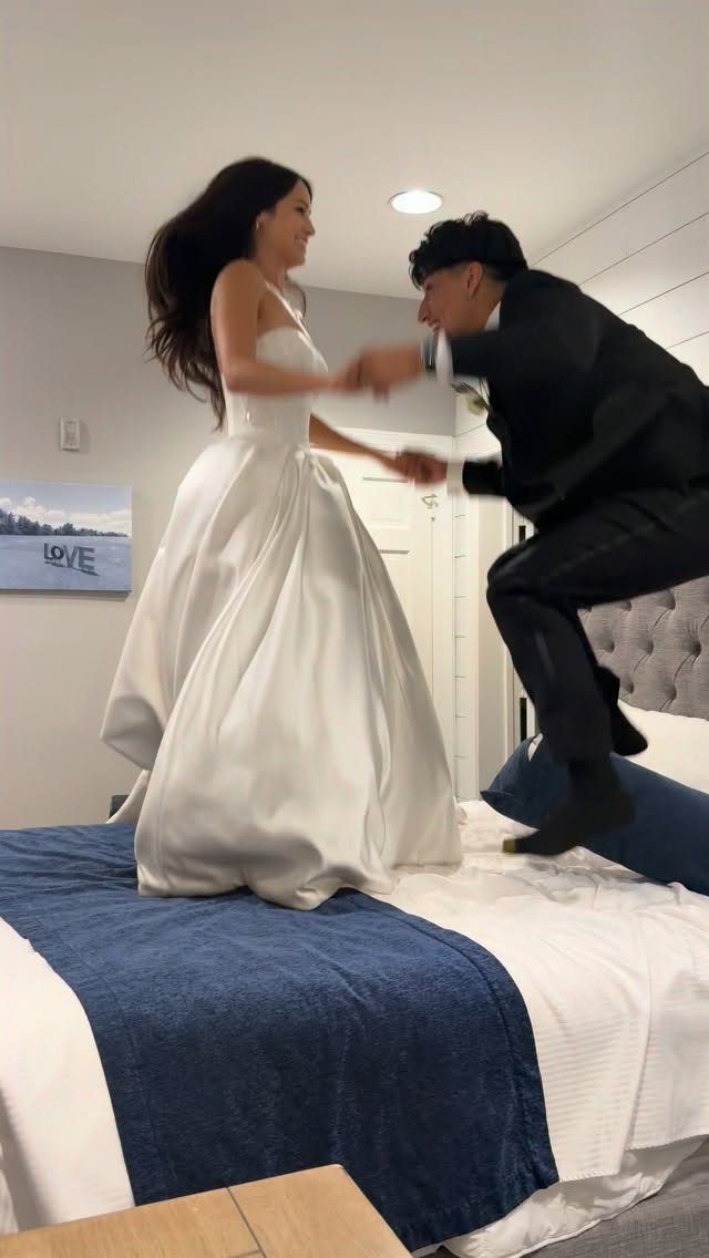 Bride and groom jumping and dancing playfully on a bed in a bedroom.