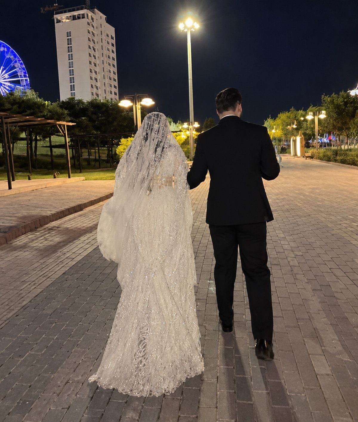Bride and groom walking away on a paved path at night near city lights.