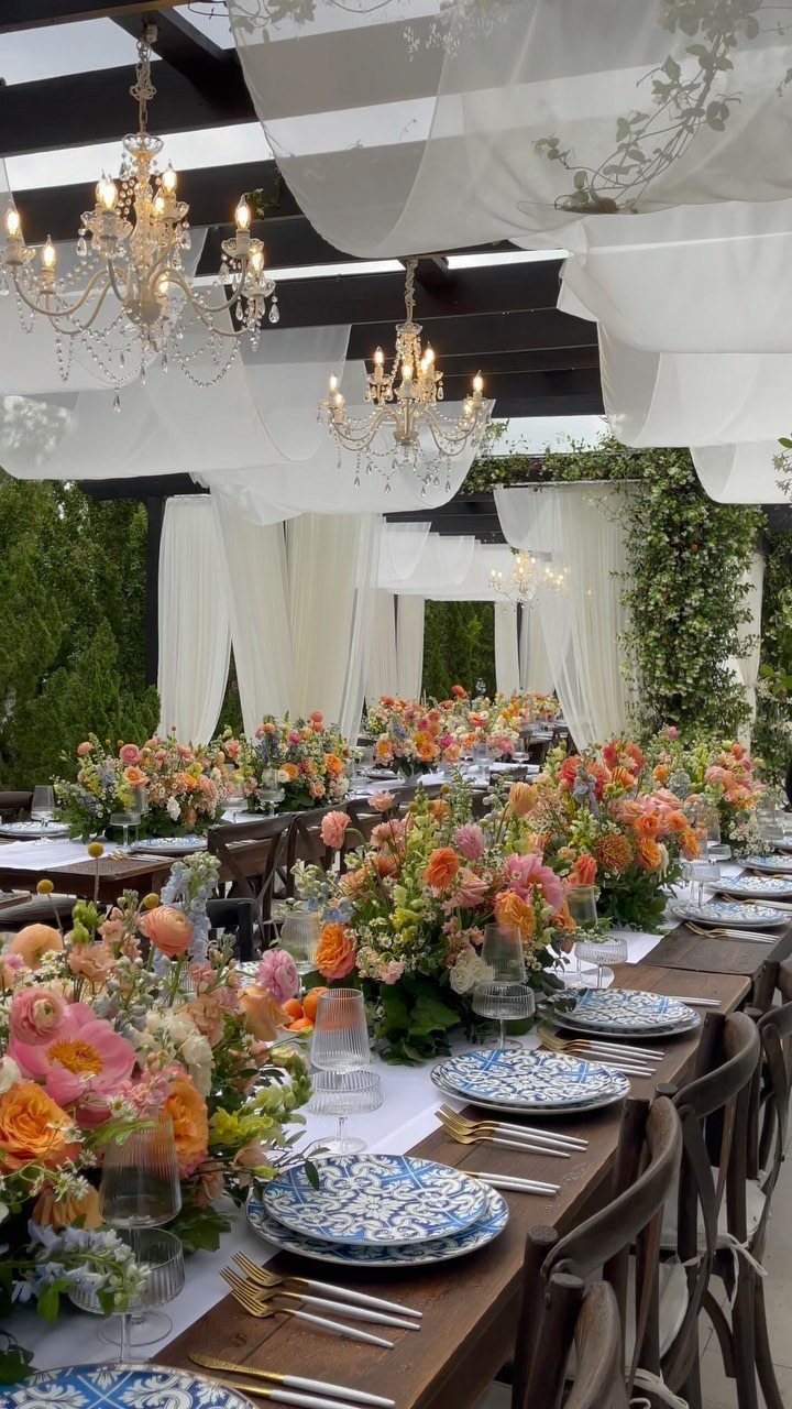 Wedding dining table under a draped pergola with chandeliers and orange floral centerpieces