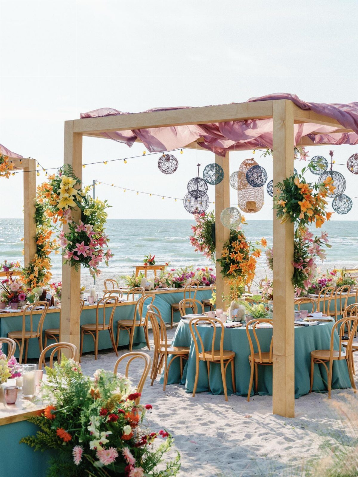 Wooden wedding pergola with purple fabric and flowers on a beach with tables.