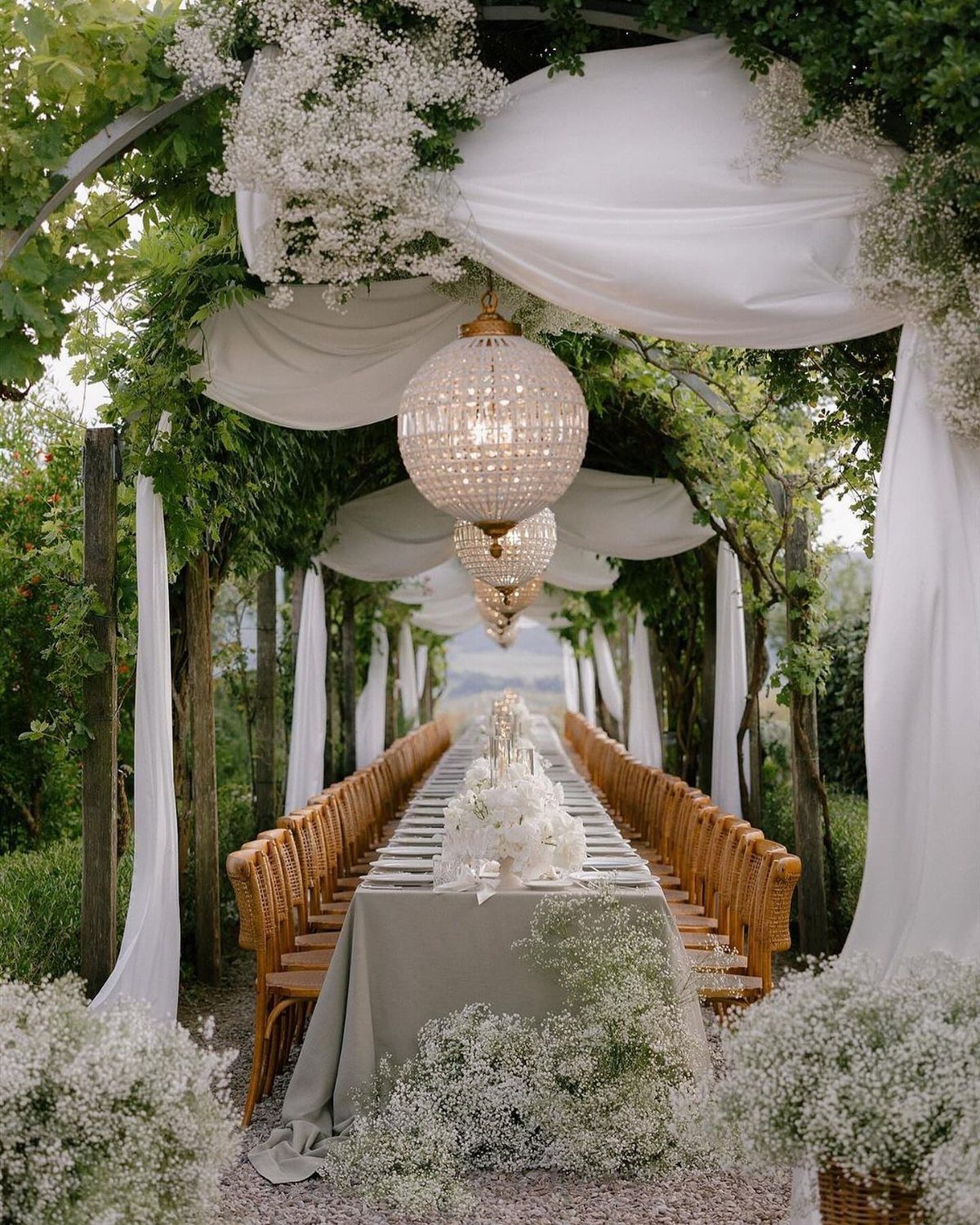 Long wedding table under a pergola with white fabric, flowers, and crystal chandeliers.