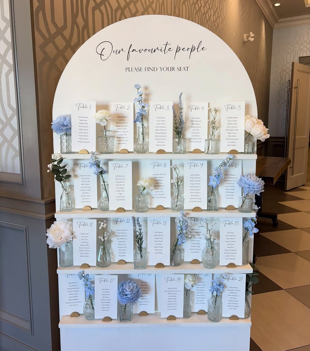 White arched seating chart with table cards and small blue and white floral arrangements.