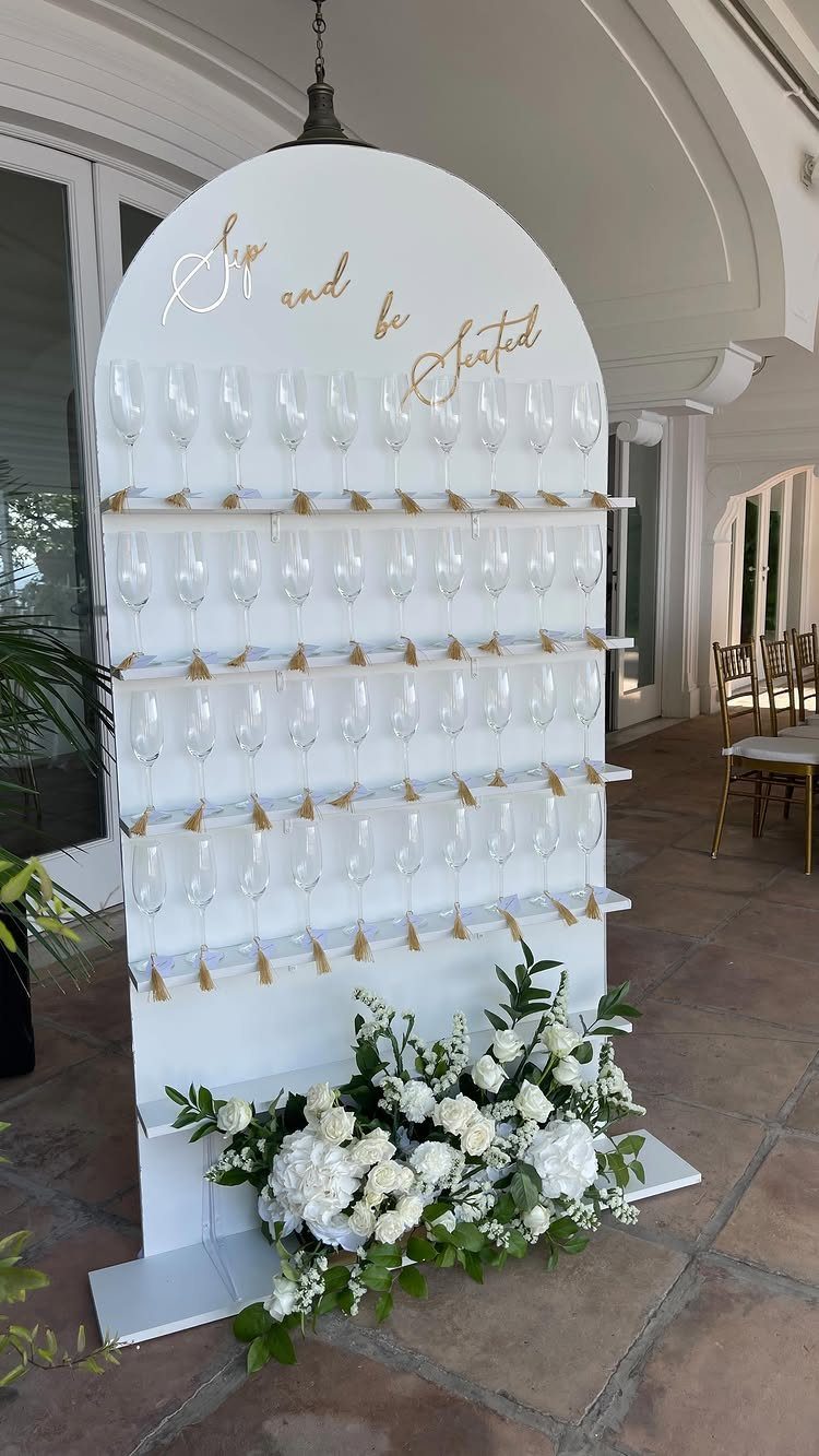 Champagne wall seating chart with white flowers and gold cursive lettering on a patio.