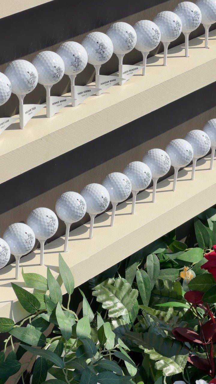 Golf balls on tees with name tags arranged on tiered shelves with greenery.