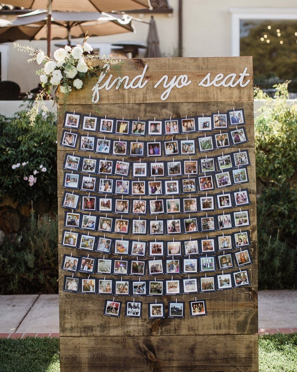 Wooden seating chart board with polaroid guest photos clipped to strings and white flowers.