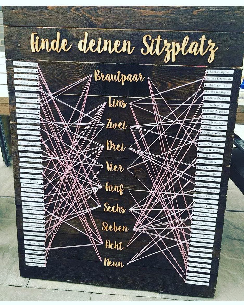 Wooden seating chart with pink strings connecting guest names to table numbers.