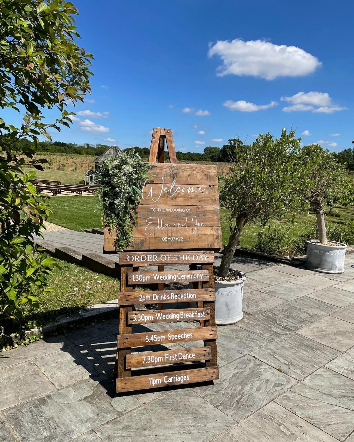Rustic wooden welcome and schedule signs on an easel at an outdoor wedding.