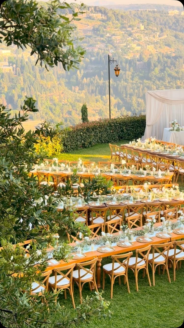 Wooden dining tables and cross-back chairs arranged on a lawn overlooking rolling hills.