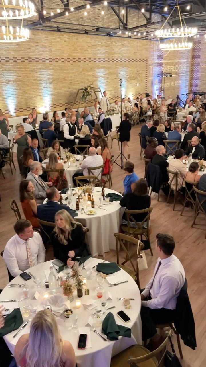 Wedding guests seated at round tables with white linens in a brick venue.