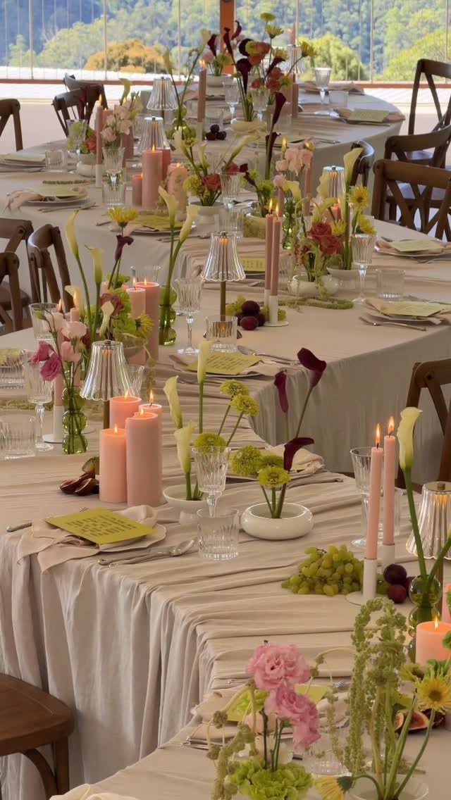 Long rectangular tables with beige linens, pink candles, and floral centerpieces outdoors.
