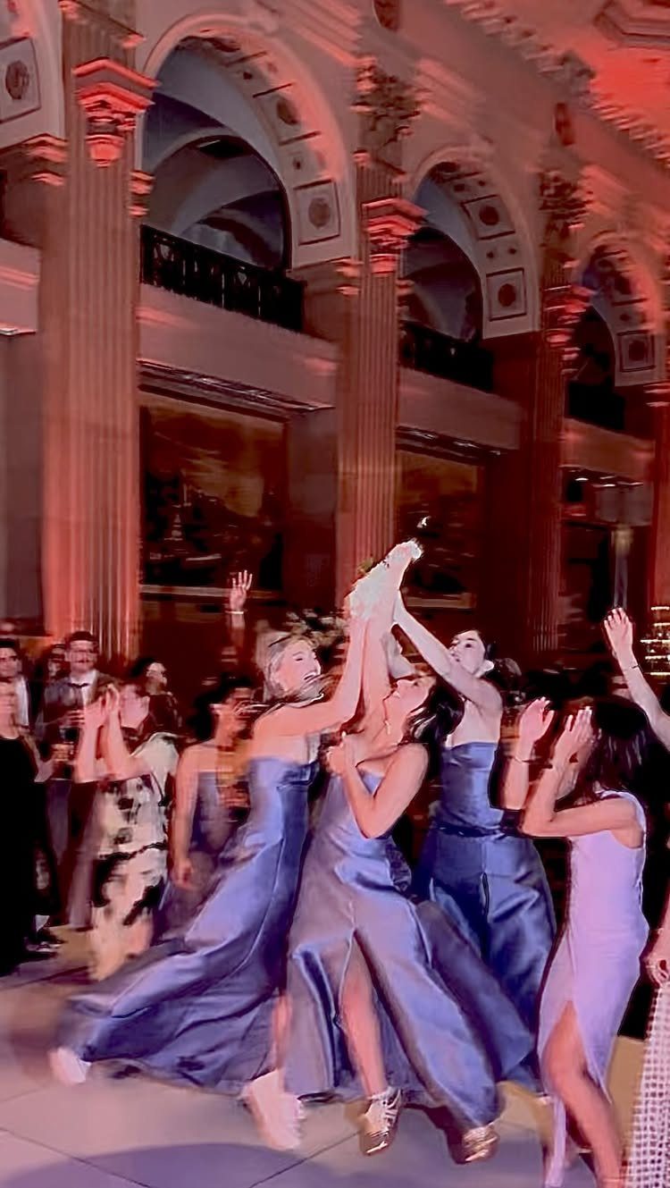 Women in formal gowns jumping to catch a white bridal bouquet in a ballroom.