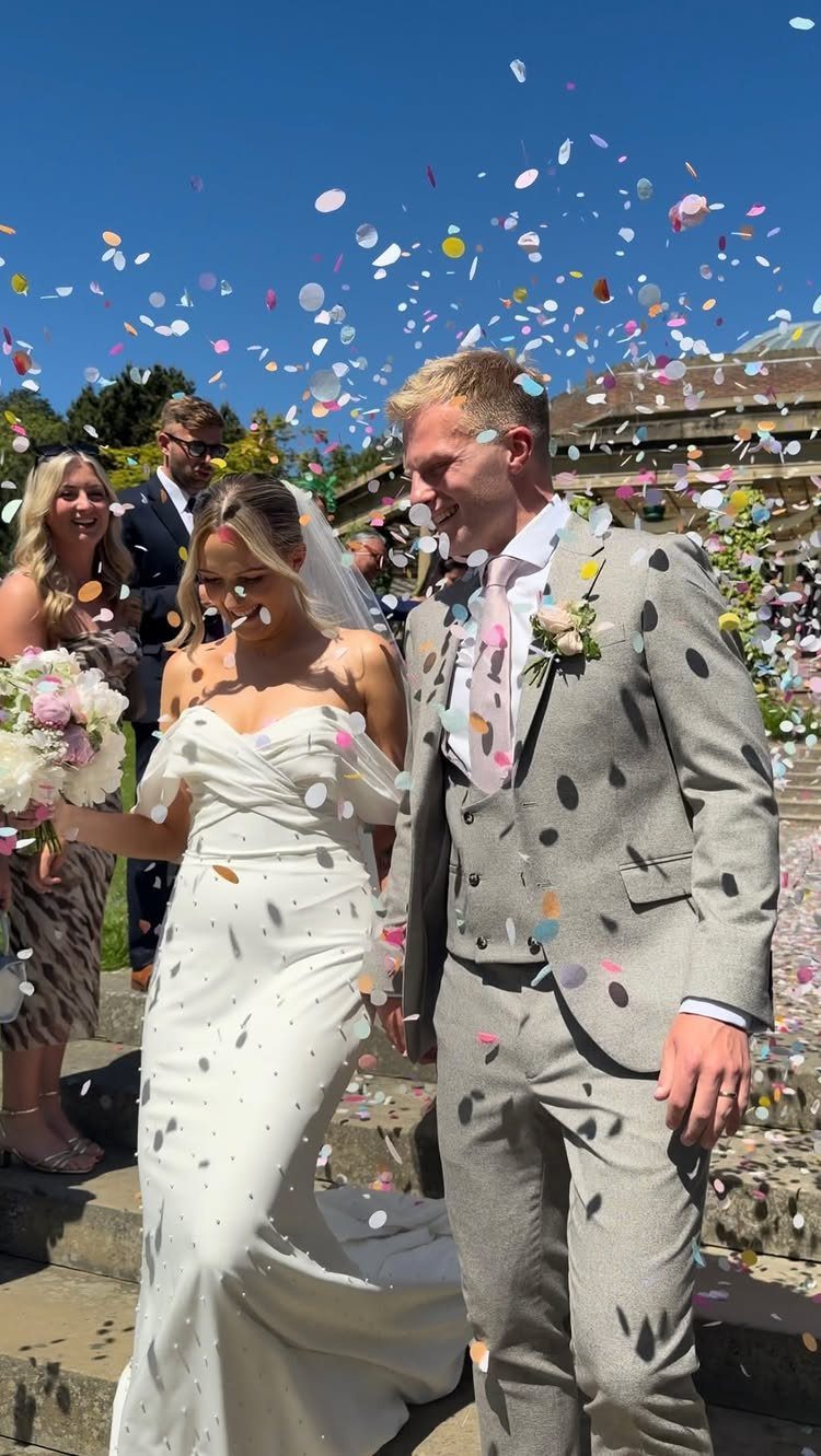 Bride and groom walking through colorful confetti thrown by guests outdoors.