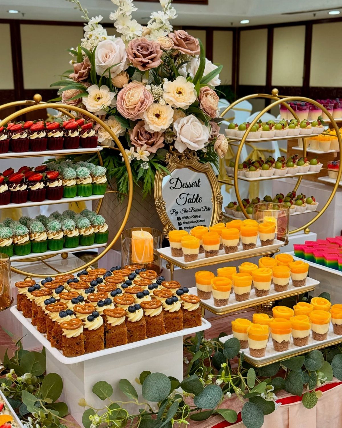 Dessert table with tiered gold stands, mini cakes, and a large floral arrangement.