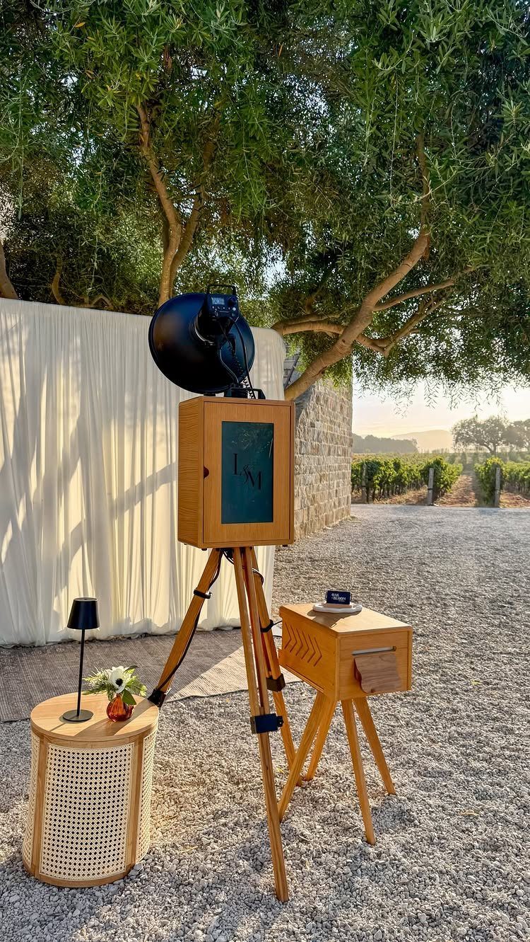 Wooden photo booth camera on a tripod with accompanying small tables outdoors.